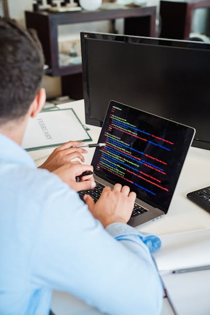 A person seated at a desk during a home relocation planning session, working on a laptop displaying lines of colorful code or data related to moving logistics. The workspace includes a black monitor, 
